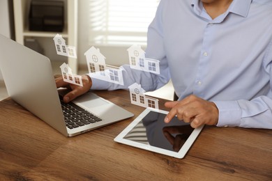 Image of Real estate agent looking for housing via laptop and tablet at wooden table, closeup. Illustrations of buildings over devices