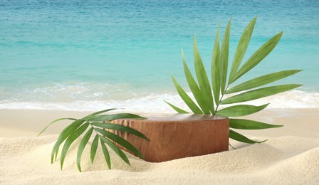 Image of Podium for product presentation with green leaves on sand near turquoise ocean, banner design. Stylish promotion poster