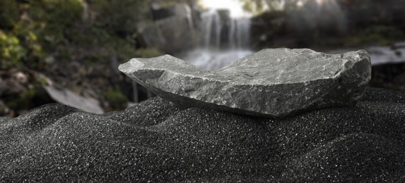 Image of Stone as podium for product presentation on black sand against waterfall, banner design. Stylish promotion poster
