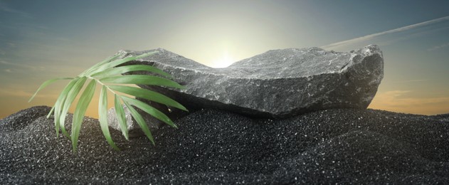 Image of Podium for product presentation. Stone and green leaf on black sand under sky, banner design. Stylish promotion poster
