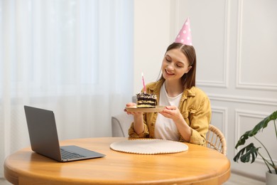 Photo of Birthday celebration. Happy woman with piece of cake having video call via laptop at home