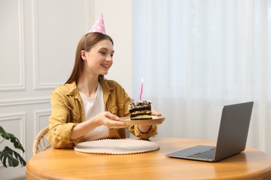 Photo of Birthday celebration. Happy woman with piece of cake having video call via laptop at home
