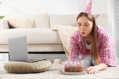 Photo of Birthday celebration. Woman blowing candles on cake while having video call via laptop at home