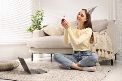 Photo of Birthday celebration. Happy woman with cupcake having video call via laptop at home
