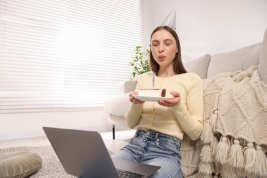 Photo of Birthday celebration. Woman blowing candle on piece of cake while having video call via laptop at home