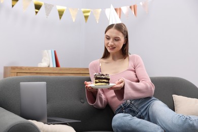 Photo of Birthday celebration. Happy woman with piece of cake having video call via laptop on sofa at home