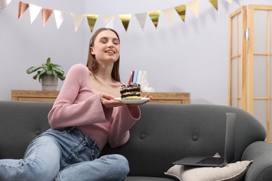 Photo of Birthday celebration. Happy woman with piece of cake having video call via laptop on sofa at home