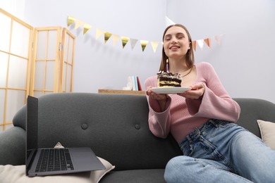Photo of Birthday celebration. Happy woman with piece of cake having video call via laptop on sofa at home