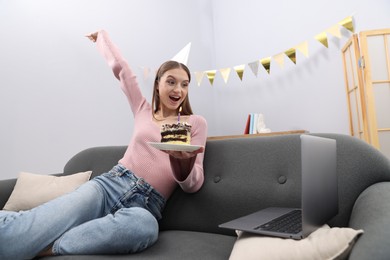 Photo of Birthday celebration. Happy woman with piece of cake having video call via laptop on sofa at home