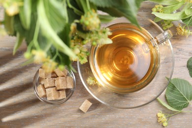 Photo of Tasty linden tea in cup, sugar cubes, leaves and flowers on wooden table, flat lay