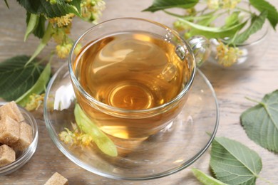Photo of Tasty linden tea in cup, sugar cubes, leaves and flowers on wooden table, closeup