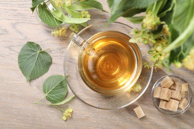 Photo of Tasty linden tea in cup, sugar cubes, leaves and flowers on wooden table, flat lay