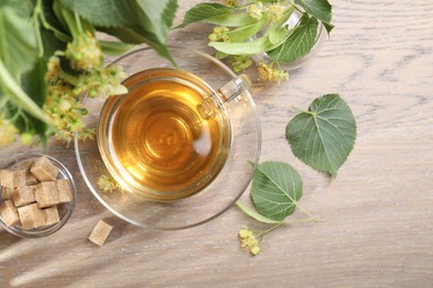 Photo of Tasty linden tea in cup, sugar cubes, leaves and flowers on wooden table, flat lay