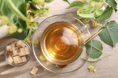 Photo of Tasty linden tea in cup, sugar cubes, leaves and flowers on wooden table, flat lay