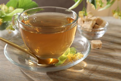 Photo of Tasty linden tea in cup, spoon, leaves and flowers on wooden table, closeup