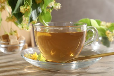 Photo of Tasty linden tea in cup, spoon, leaves and flowers on wooden table against grey background, closeup