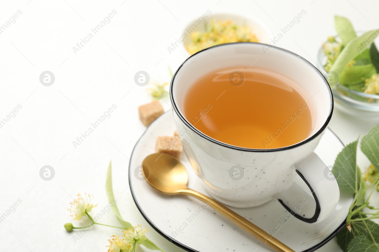Tasty linden tea in cup, spoon, sugar cubes, leaves and flowers on white table, closeup. Space for text Photo of Tasty linden tea in cup, spoon, sugar cubes, leaves and flowers on white table, closeup. Space for text