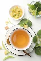 Photo of Tasty linden tea in cup, spoon, sugar cubes, leaves and flowers on white table, flat lay