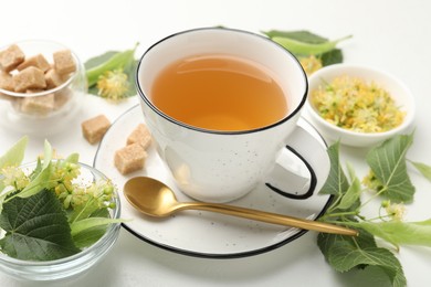 Photo of Tasty linden tea in cup, spoon, sugar cubes, leaves and flowers on white table, closeup