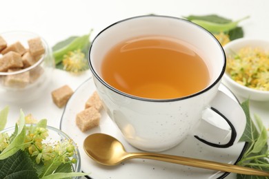 Photo of Tasty linden tea in cup, spoon, sugar cubes, leaves and flowers on white table, closeup