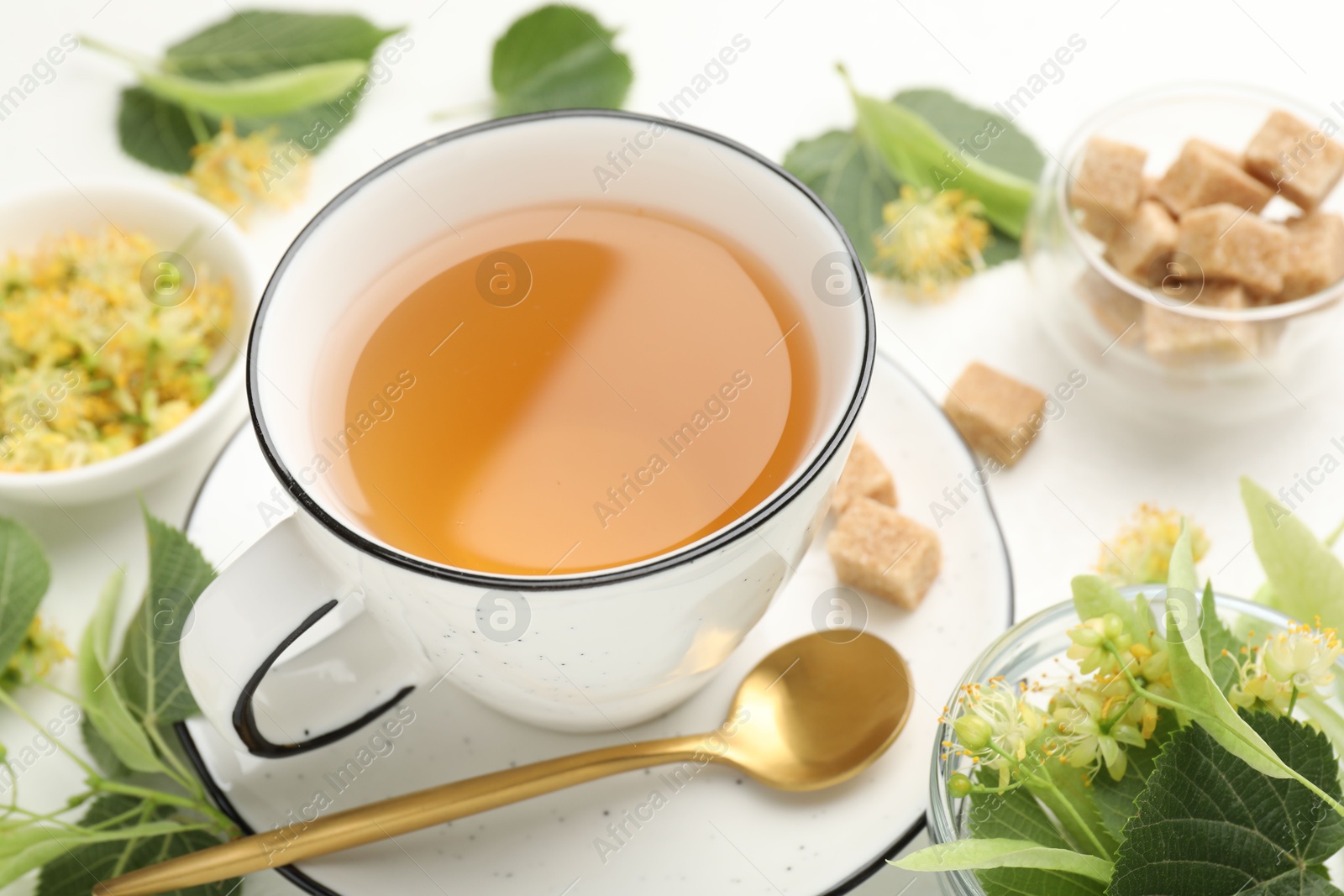 Tasty linden tea in cup, spoon, sugar cubes, leaves and flowers on white table, closeup Photo of Tasty linden tea in cup, spoon, sugar cubes, leaves and flowers on white table, closeup