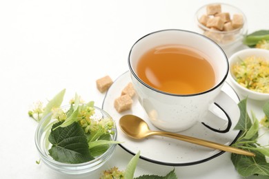 Photo of Tasty linden tea in cup, spoon, sugar cubes, leaves and flowers on white table, closeup