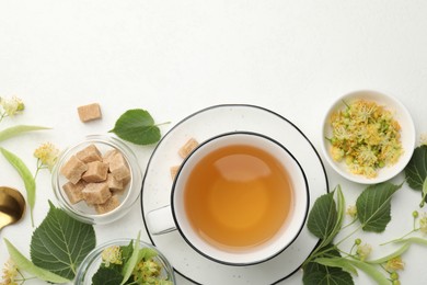 Photo of Tasty linden tea in cup, spoon, sugar cubes, leaves and flowers on white table, flat lay