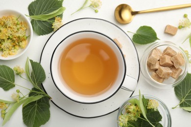 Photo of Tasty linden tea in cup, spoon, sugar cubes, leaves and flowers on white table, flat lay