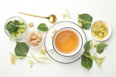 Photo of Tasty linden tea in cup, spoon, sugar cubes, leaves and flowers on white table, flat lay