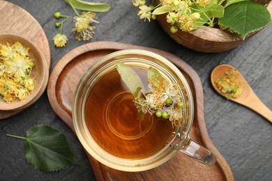 Photo of Tasty linden tea in cup, spoon, leaves and flowers on dark textured table, flat lay