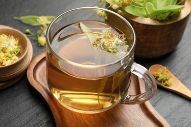 Photo of Tasty linden tea in cup, leaves and flowers on dark textured table, closeup