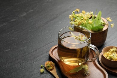 Photo of Tasty linden tea in cup, spoon, leaves and flowers on dark textured table, closeup. Space for text