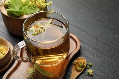 Photo of Tasty linden tea in cup, spoon, leaves and flowers on dark textured table, closeup