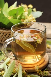 Photo of Tasty linden tea in cup, leaves and flowers on wooden table against dark background, closeup