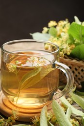 Photo of Tasty linden tea in cup, leaves and flowers on wooden table against dark background, closeup