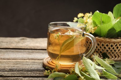 Photo of Tasty linden tea in cup, leaves and flowers on wooden table against dark background, closeup. Space for text