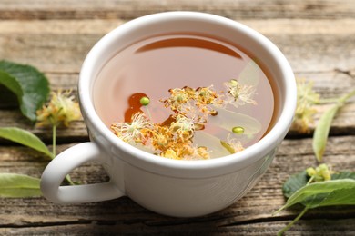 Photo of Tasty linden tea in cup, leaves and flowers on wooden table, closeup