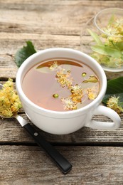 Photo of Tasty linden tea in cup, spoon, leaves and flowers on wooden table, closeup