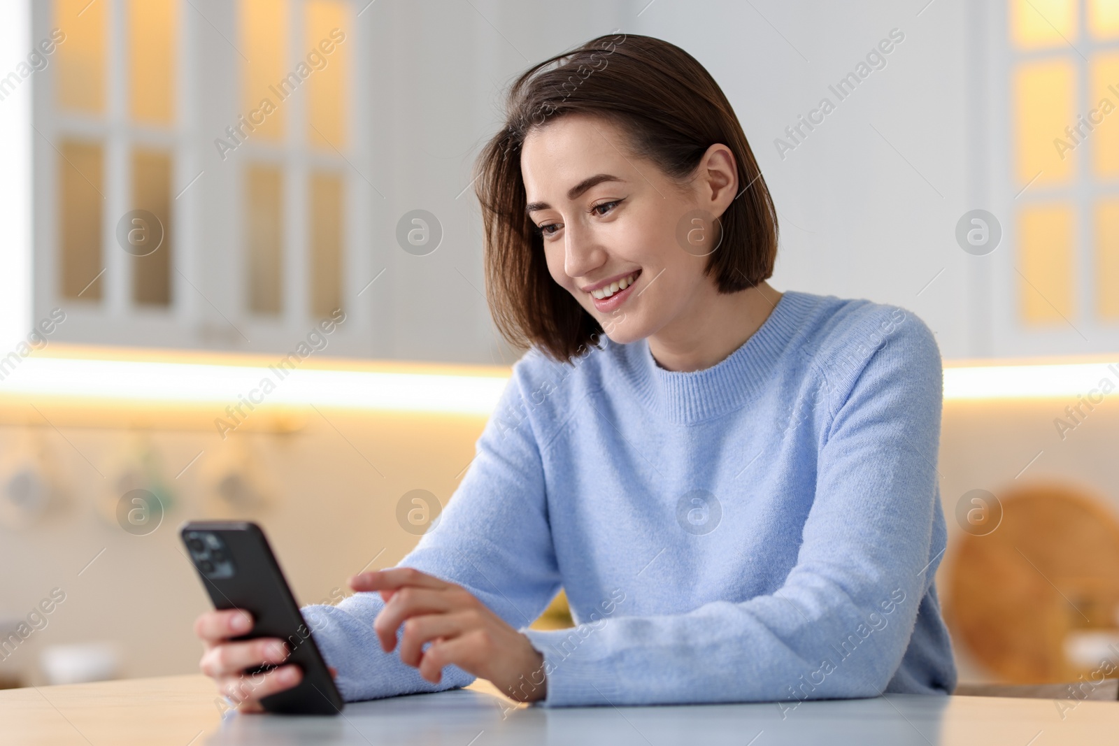 Woman using modern smartphone at table in kitchen Photo of Woman using modern smartphone at table in kitchen
