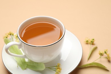 Photo of Tasty linden tea in cup, leaves and flowers on beige table, closeup