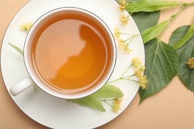 Photo of Tasty linden tea in cup, leaves and flowers on beige table, flat lay