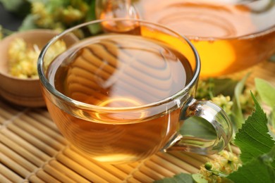 Photo of Tasty linden tea in cup, leaves and flowers on bamboo mat, closeup