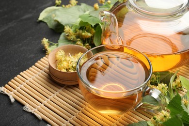 Photo of Tasty linden tea in cup, teapot, leaves and flowers on dark textured table, closeup
