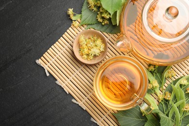 Photo of Tasty linden tea in cup, teapot, leaves and flowers on dark textured table, top view