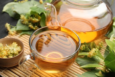 Photo of Tasty linden tea in cup, teapot, leaves and flowers on dark table, closeup