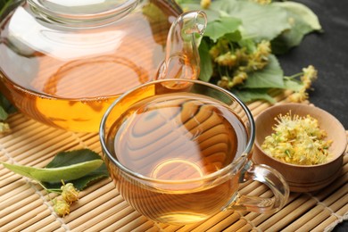 Photo of Tasty linden tea in cup, teapot, leaves and flowers on dark table, closeup