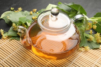 Photo of Tasty linden tea in teapot, leaves and flowers on dark textured table, closeup