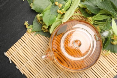 Photo of Tasty linden tea in teapot, leaves and flowers on dark textured table, flat lay