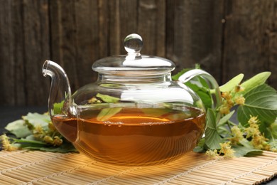 Photo of Tasty linden tea in teapot, leaves and flowers on table against wooden background, closeup