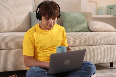 Photo of Teenage student with cup of drink using laptop while studying on floor at home